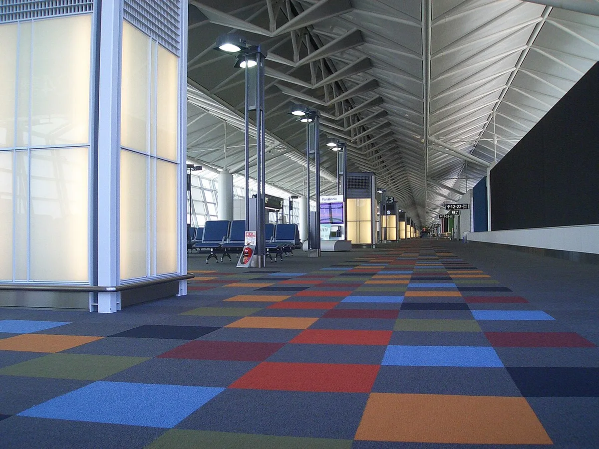 Couloir désert d’une aérogare près d’une porte d’embarquement, avec des rangées de sièges bleus vides le long d’une cloison translucide et une moquette à dalles colorées qui file vers le fond du bâtiment.